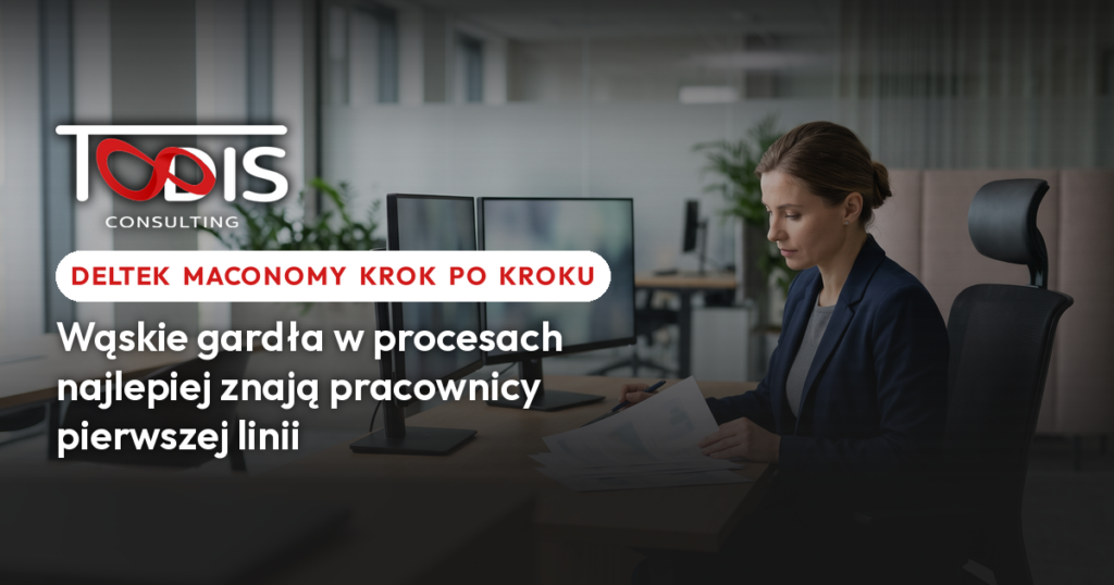 Kobieta w granatowym żakiecie przegląda wydrukowane wykresy przy biurku z dwoma monitorami w jasnym biurze